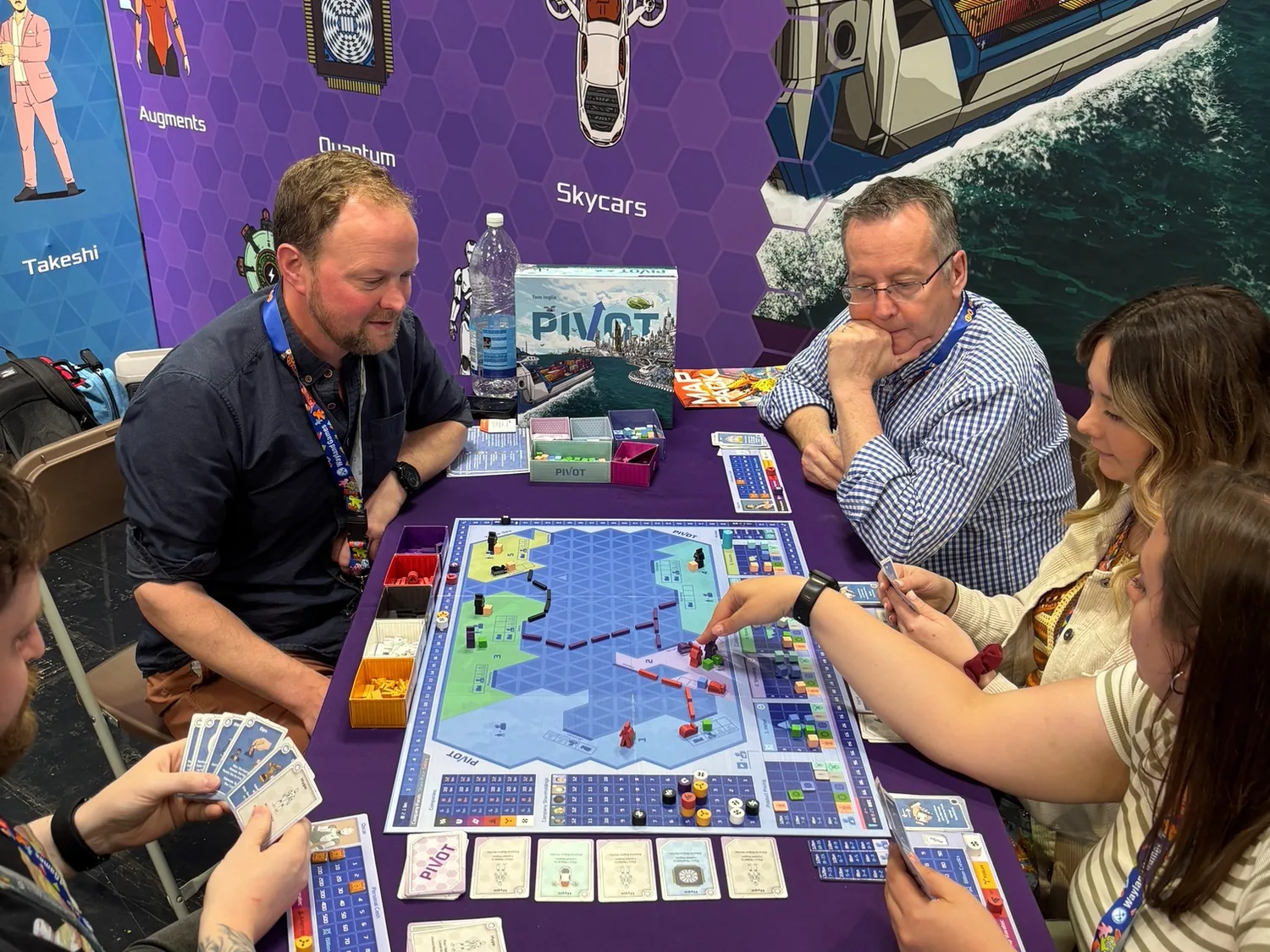 Five players around a table mid-game, the Pivot board and cards in play
